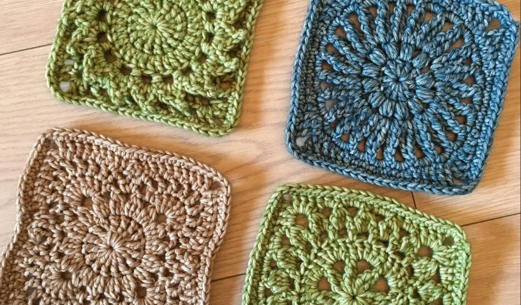 Four handmade crochet granny squares in earthy tones of green, blue, and beige, laid out on a wooden surface. Each square features intricate crochet patterns with a lacy and textured design.