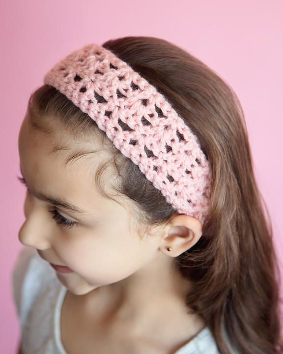 A young girl wearing a soft pink handmade crochet headband with an intricate lacy pattern. The headband is lightweight and stylish, complementing her long brown hair against a pastel pink background.