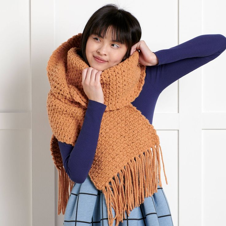 A young woman wearing a cozy, oversized handmade crochet scarf in a warm brown shade. The scarf features a textured stitch pattern and long fringe, wrapping snugly around her neck for a stylish winter look.