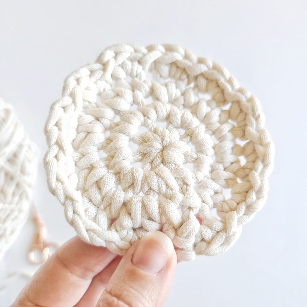 A close-up of a hand holding a round, handmade crochet face scrubbie made from soft, eco-friendly cotton yarn in a natural off-white color. The scrubbie features a textured stitch pattern, perfect for gentle exfoliation and sustainable skincare.