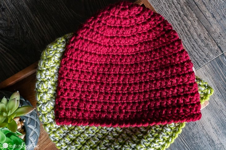 A cozy, handmade crochet beanie in a rich red color, neatly stitched in a round pattern. It is placed on top of another crochet beanie in green and gray tones, resting on a wooden surface with a small succulent plant nearby, adding a natural touch.