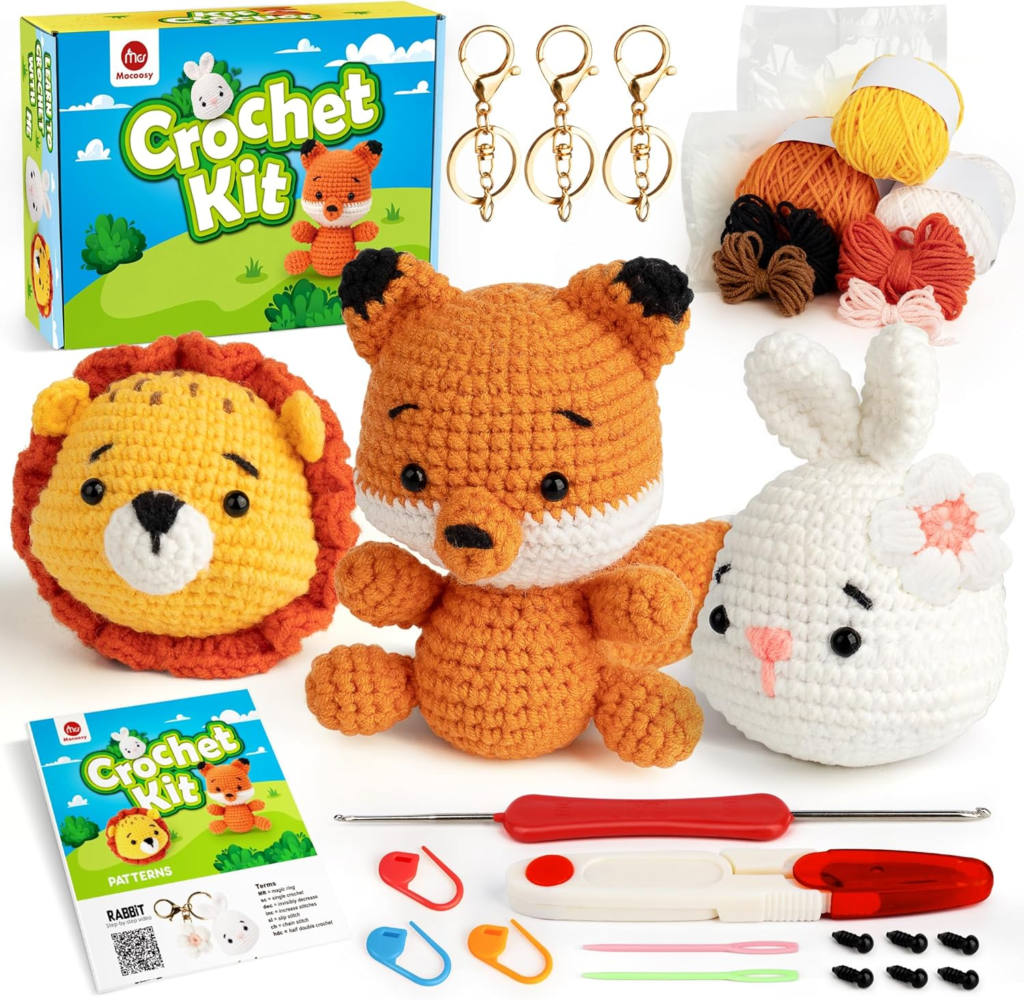 A complete crochet kit featuring materials to create three amigurumi animal keychains: a lion, a fox, and a rabbit with a flower. The kit includes colorful yarn, a red-handled crochet hook, stitch markers, embroidery needles, safety eyes, keychain rings, and an instruction booklet. The packaging is bright and playful, labeled "Crochet Kit" with cartoon-style graphics.