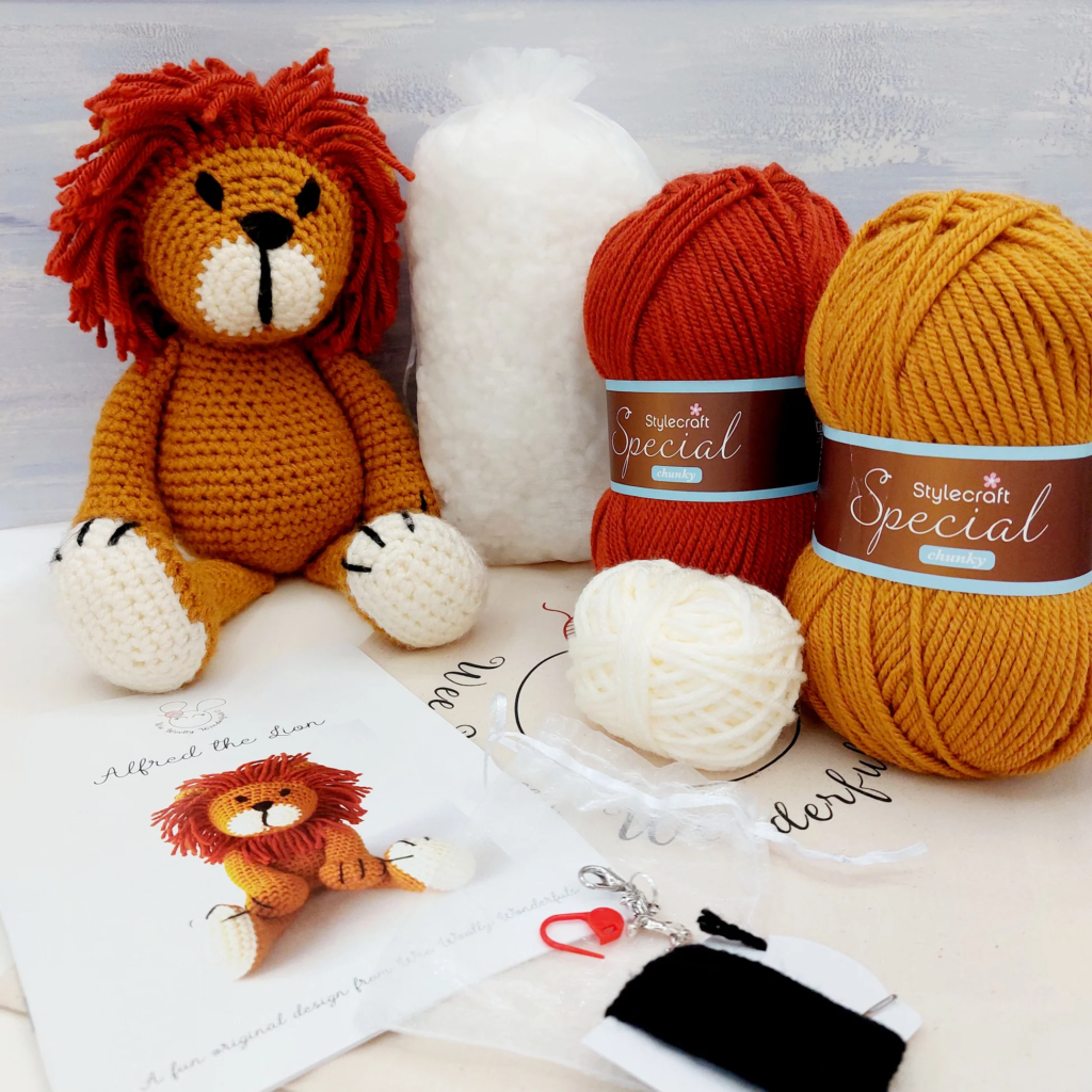 A crochet kit featuring materials to create an amigurumi lion named "Alfred the Lion." The kit includes mustard and rust-colored chunky yarn, a small white yarn ball, stuffing material, a crochet pattern booklet, a stitch marker, a keychain attachment, and embroidery thread for details. A completed crochet lion with a fluffy mane, white paws, and embroidered facial features is displayed in the background.