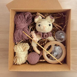 A DIY crochet kit packaged in a brown box, containing materials to create a handmade crochet baby teething ring. The kit includes beige and mauve-colored yarn, a wooden teething ring, a partially completed crochet animal head with embroidered eyes, a crochet hook, stuffing material, and a small rattle insert. The design features a soft, baby-safe amigurumi animal with a bowtie, perfect for teething and sensory play.