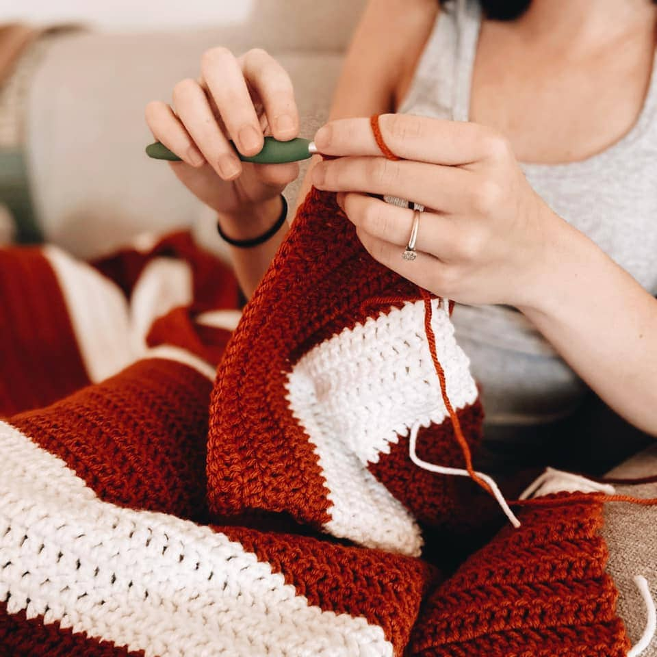 A close-up of a person crocheting a cozy blanket with a green-handled crochet hook. The blanket features a striped pattern in warm rust-red and white yarn. The individual, wearing a light gray tank top, is carefully stitching, with their hands and crochet work in focus. The soft texture of the yarn and the intricate crochet stitches highlight the craftsmanship of the handmade piece.