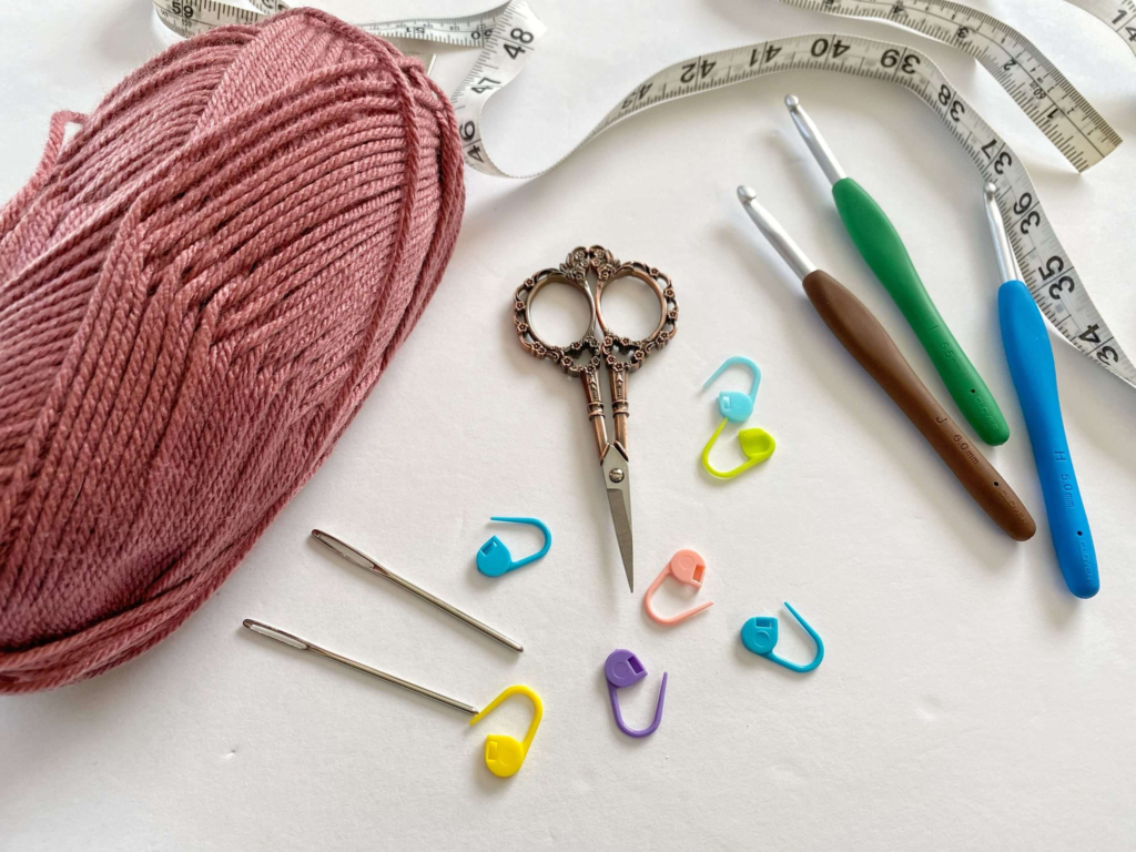 A collection of crochet tools and supplies arranged on a white surface. The set includes a skein of dusty pink yarn, three ergonomic crochet hooks in different sizes, a pair of ornate vintage-style scissors, two large sewing needles, colorful stitch markers, and a flexible measuring tape. The tools are neatly displayed, showcasing essential items for crochet projects.