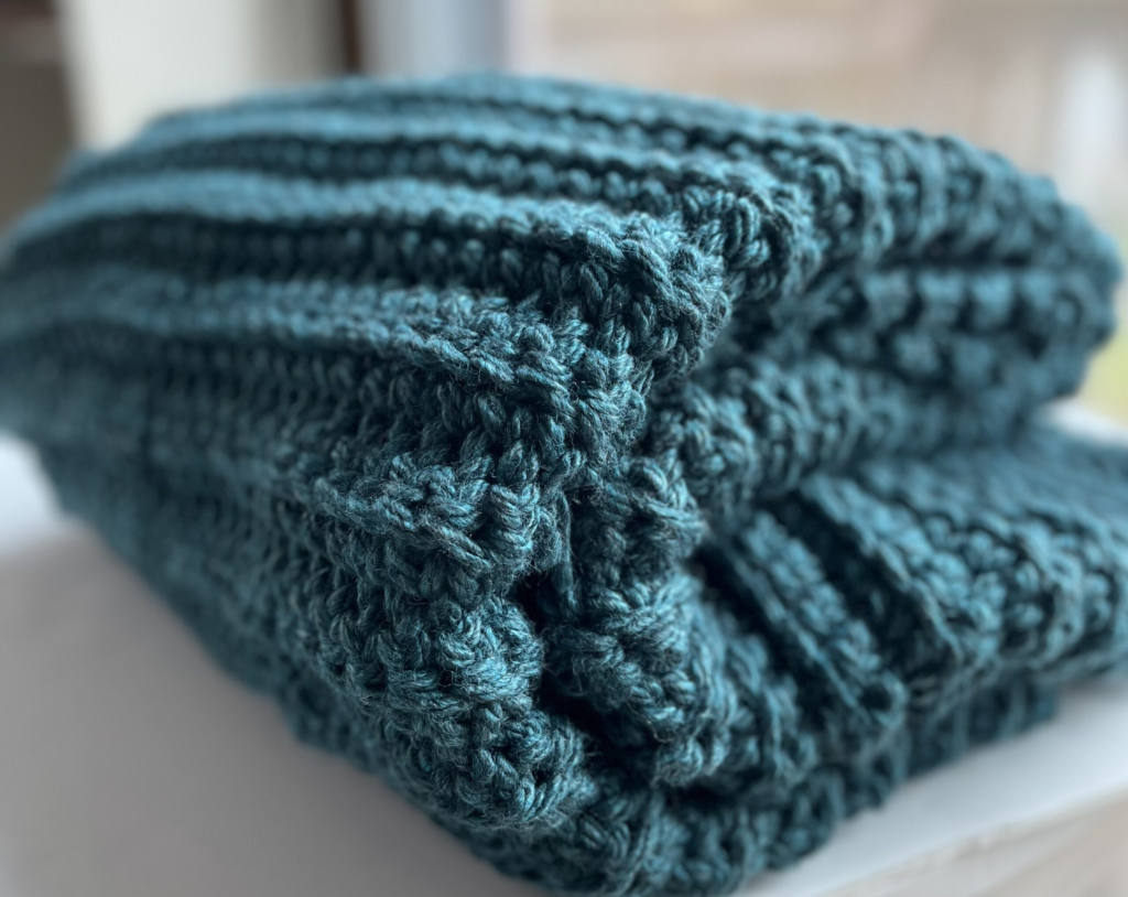 A close-up of a folded, deep teal-colored crochet blanket showcasing a beautifully textured pattern. The stitches appear raised, creating a ribbed effect. The soft and warm yarn gives the blanket a cozy appearance, making it perfect for home decor or as a thoughtful handmade gift.