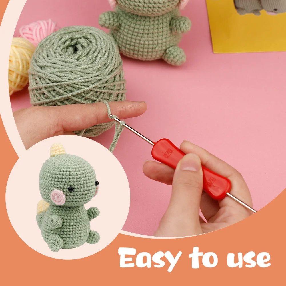 A crochet kit demonstrating an easy-to-use red-handled crochet hook with green yarn. The image features a hand crocheting and a completed green amigurumi dinosaur with pink cheeks and a yellow detail on its back. The background is pastel pink, with the text "Easy to use" in a curved orange section.